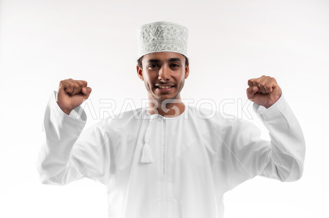 الانتصار والفوز والنجاح، مفهوم الانجاز وتحقيق الاهداف، بورتريه مقرب لشاب عربي خليجي عماني يرتدي الدشداشة والكمه ينظر الى الكاميرا يرفع يديه للاعلى بإيماءات الفرح والسرور, الشعور بالسعادة والقوة, خلفية بيضاء