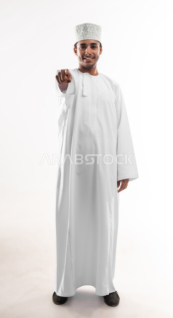 Concept of clicking, pressing and interacting with holographic elements, determining a specific location, portrait of a young Arab Gulf Omani man wearing a dishdasha and a sleeve looking at the camera raising his hand forward pointing with his index finger to a specific target, full-length body image, white background