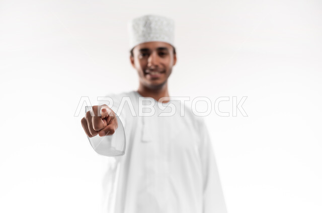 مفهوم النقر والضغط والتفاعل مع العناصر الهولوجرامية، تحديد موقع معين، بورتريه مقرب لشاب عربي خليجي عماني يرتدي الدشداشة والكمه ينظر الى الكاميرا يرفع يده للامام يشير بأصبع السبابة الى هدف محدد، خلفية بيضاء