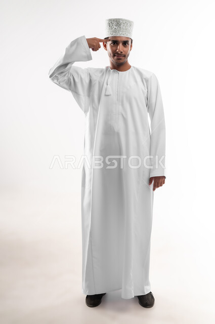 التفكير والتحليل و الذكاء والفطنه، بورتريه لرجل عربي خليجي عماني مبتسم يرتدي الدشداشة والكمه ينظر الى الكاميرا يضع إصبعه السبابة على رأسه بإيماءات العناد، مفهوم الإصرار على الرأي وفعل مايرغب، صورة الجسم بالطول الكامل, خلفية بيضاء