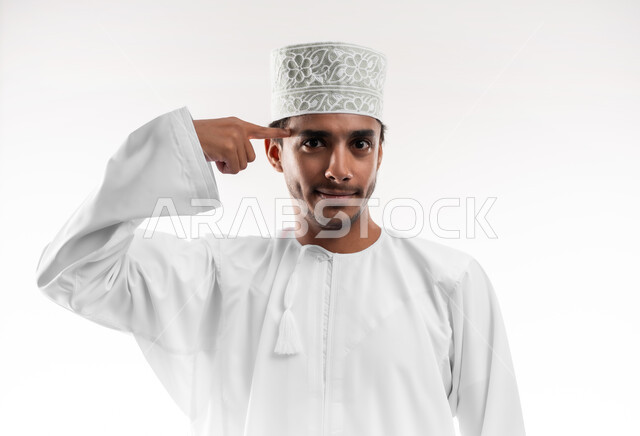 Thinking, analysis, intelligence and cleverness, close-up portrait of a smiling Omani Gulf Arab man wearing a dishdasha and a turban, looking at the camera, putting his index finger on his head with gestures of stubbornness, the concept of insistence on one's opinion and doing what one wants, white background
