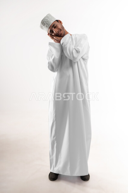 إغماض العينين والنوم بسلام، السهر لوقت متأخر من الليل، الشعور بالكسل والخمول و الارهاق، بورتريه لرجل عربي خليجي عماني مبتسم يرتدي الدشداشة والكمه يضع يديه تحت رأسه بإيماءات النعاس والتعب، صورة الجسم بالطول الكامل, خلفية بيضاء