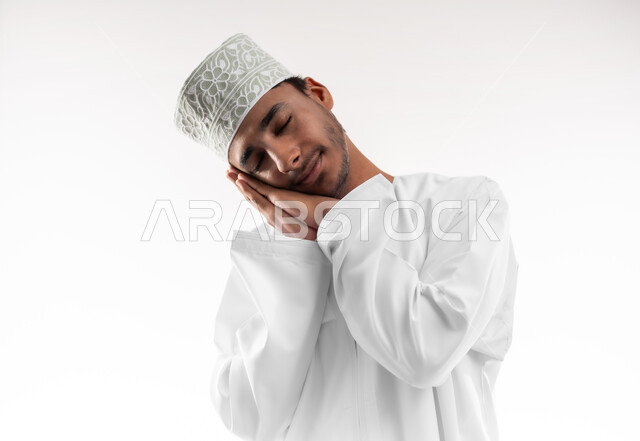 Closing eyes and sleeping peacefully, staying up late at night, feeling lazy, lethargic and exhausted, close-up portrait of a smiling Omani Gulf Arab man wearing a dishdasha and a turban, putting his hands under his head with gestures of sleepiness and fatigue, white background