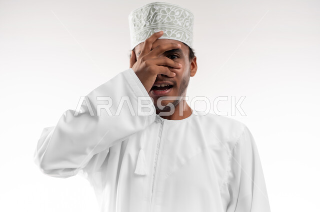 تعابير الخجل والحياء، إيماءات تذكر شيء ما بشكل مفاجئ، بورتريه مقرب لرجل عربي خليجي عماني مبتسم يرتدي الدشداشة والكمه يغطي عينيه بيده، الشعور بالفرح والسرور، خلفية بيضاء
