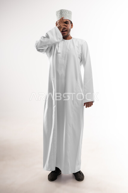 تعابير الخجل والحياء، إيماءات تذكر شيء ما بشكل مفاجئ، بورتريه لرجل عربي خليجي عماني مبتسم يرتدي الدشداشة والكمه يغطي عينيه بيده، الشعور بالفرح والسرور، صورة الجسم بالطول الكامل, خلفية بيضاء