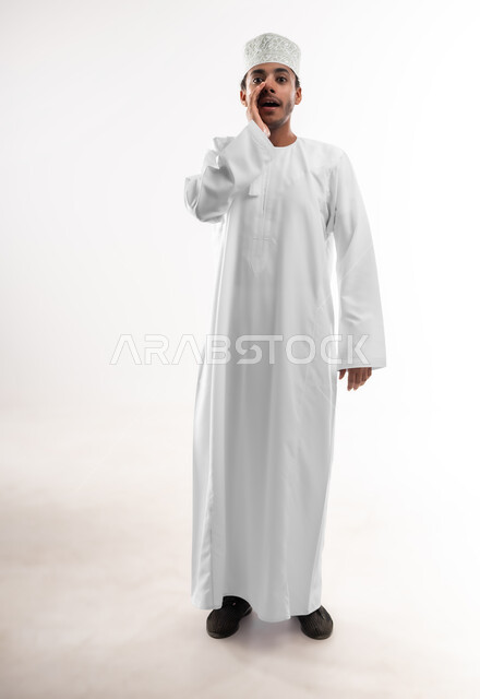 مفهوم مشاركة الأسرار، الهمس والتحدث بصوت منخفض, بورتريه لشاب عربي خليجي عماني مبتسم يرتدي الدشداشة والكمه ينظر الى الكاميرا يضع يده امام فمه بإيماءات المناداه، صورة الجسم بالطول الكامل, خلفية بيضاء