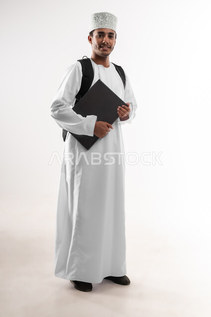 الجد والاجتهاد للوصول إلى النجاح والتفوق، بورتريه مقرب لشاب عربي خليجي عماني مبتسم يرتدي الدشداشة والكمه حمل حقيبة الظهر والكتب ينظر الى الكاميرا بإيماءات النشاط والسرور، صورة الجسم بالطول الكامل, خلفية بيضاء