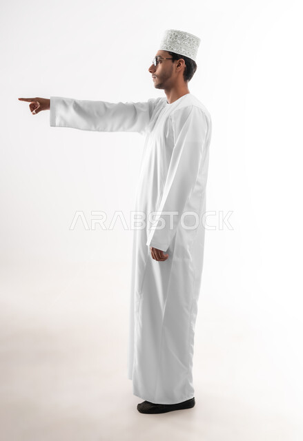 تحديد موقع معين، بورتريه مقرب لشاب عربي خليجي عماني يرتدي الدشداشة والكمه والنظارات الطبية يرفع يده للاعلى يشير بأصبع السبابة الى هدف محدد، مفهوم النقر والضغط والتفاعل مع العناصر الهولوجرامية، خلفية بيضاء
