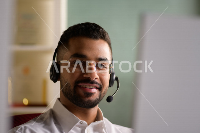 موظف عربي خليجي سعودي يعمل في خدمة العملاء ويرتدي سماعة الاتصال، استعلامات واستفسارات، الاجابه على اسئلة العملاء، اجراء اتصال فيديو من خلال الحاسوب، نظارة طبية، متابعة وتطوير العمل