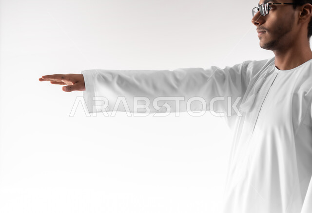 تقدير وقياس ارتفاع الأشياء، مفهوم تقديم وعرض عنصر معين، بورتريه مقرب لشاب عربي خليجي عماني يرتدي الدشداشة والكمه والنظارات الطبية يرفع يده للاعلى يحاول لمس العناصر الهولوجرامية، خلفية بيضاء