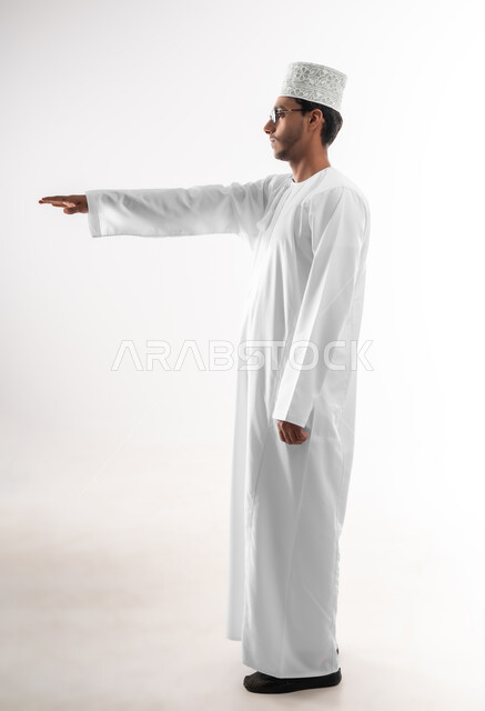 Estimating and measuring the height of objects, the concept of presenting and displaying a certain item, a portrait of a young Arab Gulf Omani man wearing a dishdasha, a sleeve and eyeglasses raising his hand up trying to touch the holographic elements, a full-length body image, white background