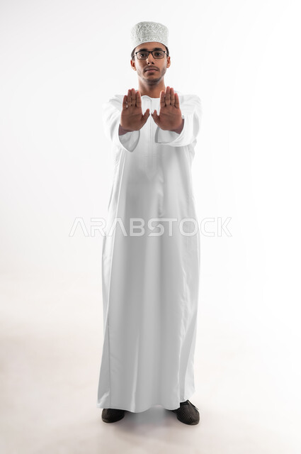 مفهوم الرفض والامتناع والانزعاج, طلب التوقف وعدم الاقتراب, بورتريه مقرب لشاب عربي خليجي عماني يرتدي الدشداشة والكمه والنظارات الطبية ينظر الى الكاميرا يمد يديه نحو الأمام مع رفع الكف بتعابير النهي عن فعل امر ما, صورة الجسم بالطول الكامل، خلفية بيضاء