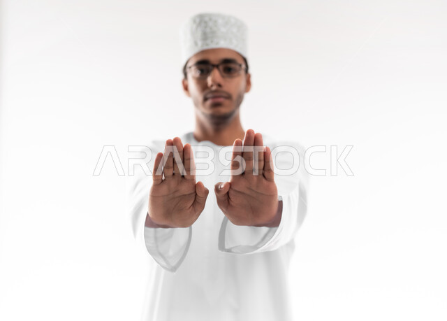 مفهوم الرفض والامتناع والانزعاج, ايماءات الحزم والجدية, طلب التوقف وعدم الاقتراب, بورتريه مقرب لشاب عربي خليجي عماني يرتدي الدشداشة والكمه والنظارات الطبية ينظر الى الكاميرا يمد يديه نحو الأمام مع رفع الكف بتعابير النهي عن فعل امر ما, خلفية بيضاء