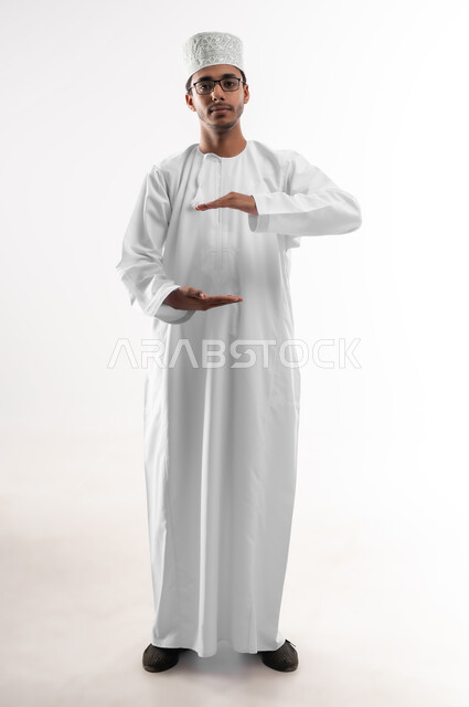 إحتواء وإمساك عنصر ما، مفهوم الاناقة و الاهتمام بالمظهر الخارجي, بورتريه لشاب عربي خليجي عماني يرتدي الدشداشة والكمه والنظارة الطبية يفتح يديه لعرض عناصر هولوجرامية ثلاثية الأبعاد، تحديد قياس وحجم شيء معين، صورة الجسم بالطول الكامل، خلفية بيضاء