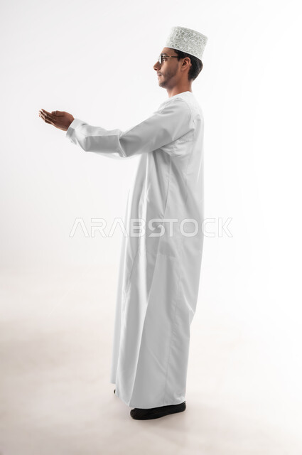 الاجتهاد في العبادة والتقرب إلى رب العالمين، بورتريه لشاب عربي خليجي عماني يرتدي الدشداشة و الكمه والنظارة الطبية يرفع يديه ويدعو الله بإيماءات التوسل والتضرع، الرغبة في تحقيق الأمنيات، صورة الجسم بالطول الكامل، خلفية بيضاء