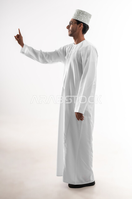 تعابير تدل على الرقم واحد, بورتريه لشاب عربي خليجي عماني يرتدي الدشداشة و الكمه والنظارات الطبية يرفع اصبع السبابة الى الاعلى بإيماءات التحذير والتنبيه، صورة الجسم بالطول الكامل، خلفية بيضاء