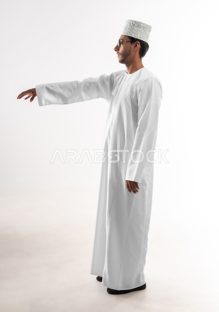 محاولة الإحاطة والإمساك بشيء ما، الإشارة إلى عنصر ما، بورتريه لشاب عربي خليجي عماني يرتدي الدشداشة و الكمه والنظارات الطبية يمد يده للأمام يحاول التقاط عناصر هولوجرامية ثلاثية الأبعاد، صورة الجسم بالطول الكامل، خلفية بيضاء