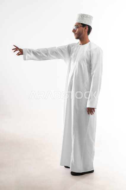 محاولة الإحاطة والإمساك بشيء ما، الإشارة إلى عنصر ما، بورتريه لشاب عربي خليجي عماني يرتدي الدشداشة و الكمه والنظارات الطبية يمد يده للأمام يحاول التقاط عناصر هولوجرامية ثلاثية الأبعاد، صورة الجسم بالطول الكامل، خلفية بيضاء