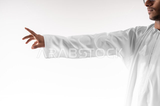 الإشارة إلى عنصر ما، بورتريه مقرب لشاب عربي خليجي عماني يرتدي الدشداشة يمد يده للأمام يحاول التقاط عناصر هولوجرامية ثلاثية الأبعاد، محاولة الإحاطة والإمساك بشيء ما، خلفية بيضاء