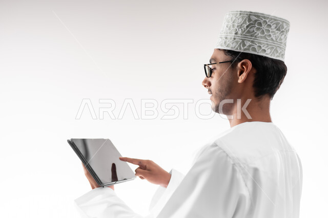 استخدام حاسة اللمس بالضغط والنقر، تصفح مواقع التواصل الاجتماعي، بورتريه مقرب لشاب عربي خليجي عماني يرتدي الدشداشة و الكمه والنظارات الطبية يحمل بيده جهاز التابلت اللوحي، الدردشة والتواصل مع الأهل والأصدقاء، خلفية بيضاء