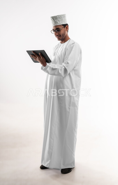 Using advanced technology, browsing social media, portrait of a young Arab Gulf Omani man wearing a dishdasha, a cufflink and glasses, holding a tablet, chatting and communicating with family and friends, full-length body image, white background