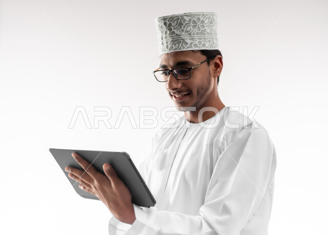 استخدام التكنولوجيا المتطورة، تصفح مواقع التواصل الاجتماعي، بورتريه مقرب لشاب عربي خليجي عماني يرتدي الدشداشة و الكمه والنظارات الطبية يحمل بيده جهاز التابلت اللوحي، الدردشة والتواصل مع الأهل والأصدقاء، خلفية بيضاء