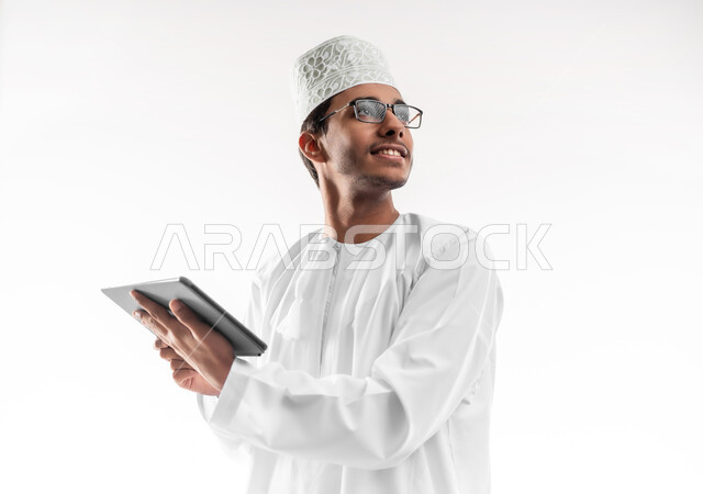 تصفح مواقع التواصل الاجتماعي، استخدام التكنولوجيا المتطورة، بورتريه مقرب لشاب عربي خليجي عماني يرتدي الدشداشة و الكمه والنظارات الطبية يحمل بيده جهاز التابلت اللوحي ينظر الى شيء ما بإيماءات السرور، الدردشة والتواصل مع الأهل والأصدقاء، خلفية بيضاء