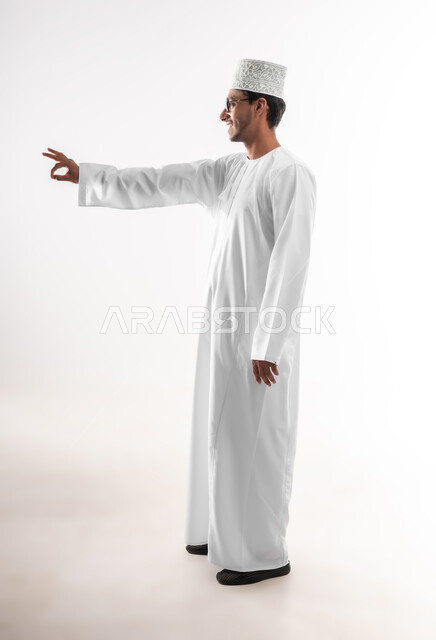 علامة الجوده و الإمتياز والتميز, مفهوم القبول والموافقه، بورتريه لشاب عربي خليجي عماني مبتسم يرتدي الدشداشة والكمه والنظارات الطبية يرفع يديه للاعلى بإيماءات الإعجاب والرضا، صورة الجسم بالطول الكامل, خلفية بيضاء