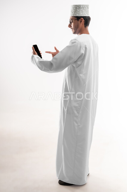 إجراء بث مباشر، التواصل و الدردشة مع الاصدقاء بإيماءات السعادة، بورتريه لشاب عربي خليجي عماني يرتدي الدشداشة والكمه والنظارات الطبية يمسك بيده الهاتف المحمول يشير إليه بإصبع السبابة يقوم بإجراء مكالمات الفيديو، صورة الجسم بالطول الكامل، خلفية بيضاء