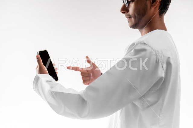 إجراء بث مباشر، التواصل و الدردشة مع الاصدقاء بإيماءات السعادة، بورتريه مقرب لشاب عربي خليجي عماني يرتدي الدشداشة والكمه والنظارات الطبية يمسك بيده الهاتف المحمول يشير إليه بإصبع السبابة يقوم بإجراء مكالمات الفيديو، خلفية بيضاء