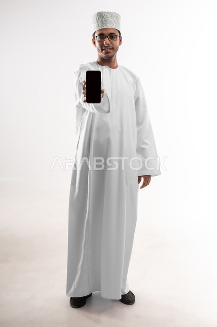 النظر الى الكاميرا بايماءات السرور والسعادة، استخدام الأجهزة التقنية الحديثة المتطورة، بورتريه لشاب عربي خليجي عماني يرتدي الدشداشة والكمه والنظارات الطبية يمسك بيده الهاتف المحمول بشاشة سوداء فارغة، صورة الجسم الطول الكامل، خلفية بيضاء
