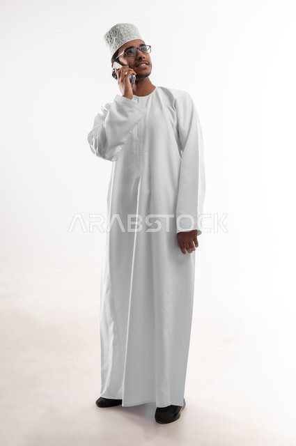 التواصل و الدردشة مع الاصدقاء بإيماءات السعادة، بورتريه مقرب لشاب عربي خليجي عماني يرتدي الدشداشة والكمه والنظارات الطبية يمسك بيده الهاتف المحمول يقوم بإجراء المكالمات الهاتفية، استخدام الأجهزة والتقنيات الحديثة، صورة الجسم بالطول الكامل، خلفية بيضاء