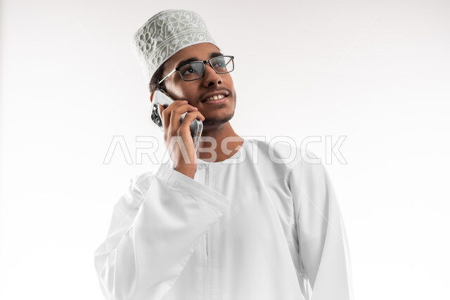 الدردشة والتواصل مع الاصدقاء بإيماءات السعادة والفرح، بورتريه مقرب لشاب عربي خليجي عماني يرتدي الدشداشة والكمه والنظارات الطبية يمسك بيده الهاتف المحمول يقوم بإجراء المكالمات الهاتفية، استخدام الأجهزة والتقنيات الحديثة، خلفية بيضاء