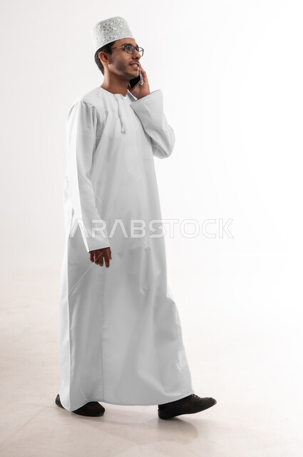 التواصل و الدردشة مع الاصدقاء بإيماءات السعادة، بورتريه مقرب لشاب عربي خليجي عماني يرتدي الدشداشة والكمه والنظارات الطبية يمسك بيده الهاتف المحمول يقوم بإجراء المكالمات الهاتفية، استخدام الأجهزة والتقنيات الحديثة، صورة الجسم بالطول الكامل، خلفية بيضاء