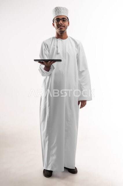The concept of e-shopping, using advanced technology, displaying applications and services of modern technical devices, a portrait of a young Arab Gulf Omani man wearing a dishdasha, a sleeve and medical glasses, looking at the camera holding a portable tablet in his hand, a full-length body image, white background