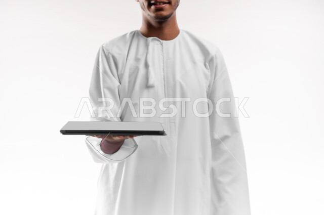 The concept of e-shopping, using advanced technology, displaying applications and services of modern technical devices, close-up portrait of a young Gulf Arab Omani man wearing a dishdasha holding a portable tablet, white background