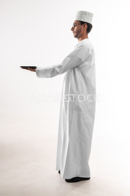 مفهوم التسوق الالكتروني، استخدام التكنولوجيا المتطورة، عرض تطبيقات وخدمات الأجهزة التقنية الحديثة، بورتريه مقرب لشاب عربي خليجي عماني يرتدي الدشداشة والكمه والنظارة الطبية  يحمل بيده جهاز التابلت المحمول، صورة الجسم الطول الكامل، خلفية بيضاء