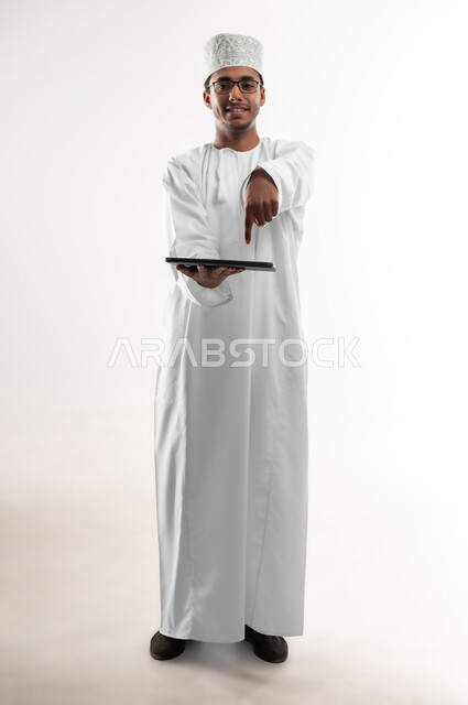 استخدام التكنولوجيا المتطورة، عرض تطبيقات وخدمات الأجهزة التقنية الحديثة، بورتريه مقرب لشاب عربي خليجي عماني يرتدي الدشداشة والكمه والنظارة الطبية  يحمل بيده جهاز التابلت المحمول يشير اليه باصبع السبابة، صورة الجسم الطول الكامل، خلفية بيضاء