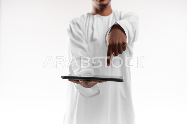 Using advanced technology, displaying applications and services of modern technical devices, close-up portrait of a young Gulf Arab Omani man wearing a dishdasha holding a portable tablet in his hand, pointing at it with his index finger, white background