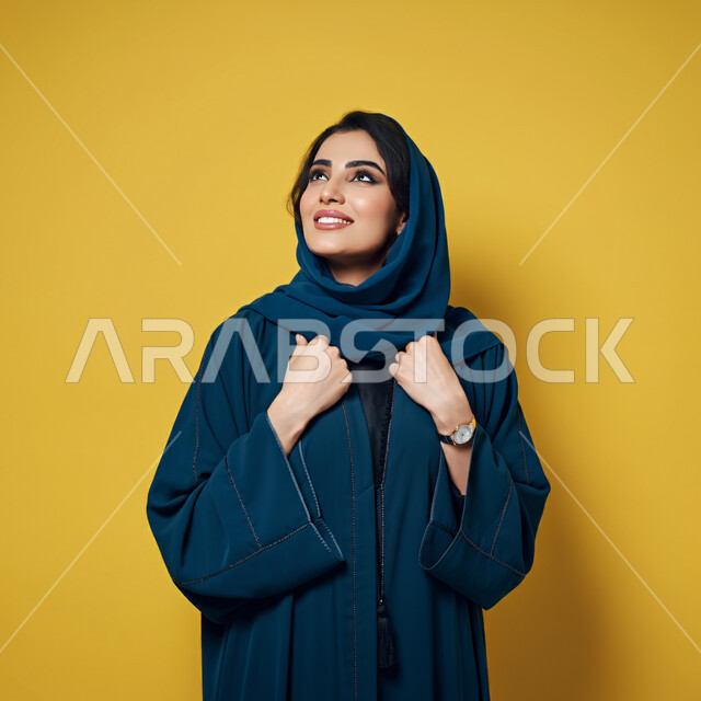Expressions of joy, pleasure and happiness, interest in elegance and appearance, concept of femininity and softness, close-up portrait of a smiling veiled Saudi Arabian Gulf woman wearing a blue abaya looking up with gestures of self-confidence and joy, yellow background