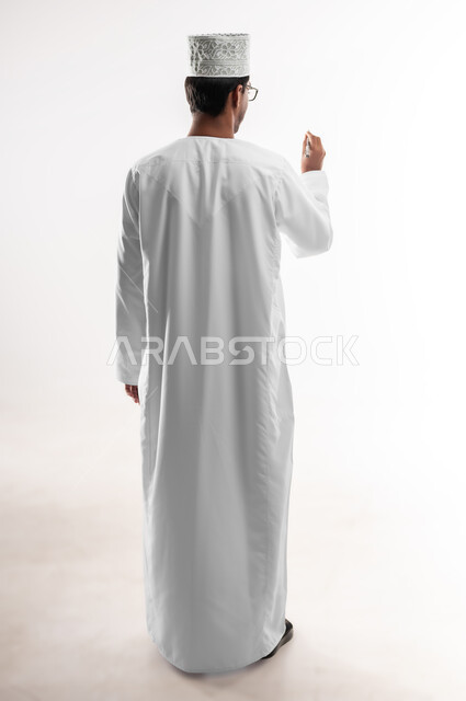 Taking notes, getting ready to write and mark something, portrait from behind of a young Arab Gulf Omani man wearing a dishdasha and a sleeve holding a pen writing something, concept of education according to the prescribed curricula, full-length body image, white background