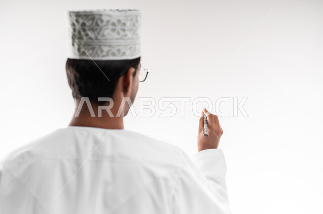 تدوين وتسجيل الملاحظات، الإستعداد للكتابة وتحديد شيء ما, بورتريه مقرب من الخلف لشاب عربي خليجي عماني يرتدي الدشداشة والكمه يمسك بيده قلم يكتب شيء ما, مفهوم التعليم بالمناهج المقررة، خلفية بيضاء