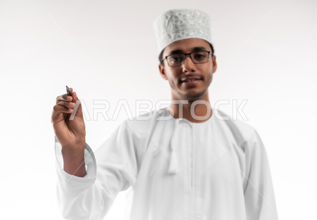 Preparing to write and outline something, taking notes, close-up portrait of a young Omani Gulf Arab man wearing a dishdasha, a sleeve and glasses looking at the camera holding a pen writing something, concept of education with prescribed curricula, white background