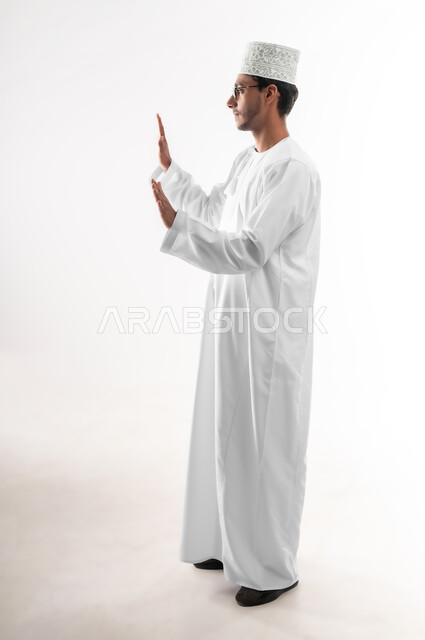 طلب التوقف والإمتناع عن فعل شيء ما، حماية النفس من ضرر خارجي, الرعب والذعر والرهبة، بورتريه لشاب عربي خليجي عماني يرتدي الدشداشة والكمه والنظارة الطبية يرفع يديه للأعلى بإيماءات الخوف والاستسلام، صورة الجسم بالطول الكامل, خلفية بيضاء