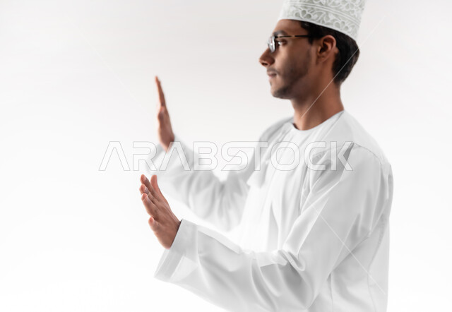 طلب التوقف والإمتناع عن فعل شيء ما، حماية النفس من ضرر خارجي, الرعب والذعر والرهبة، بورتريه مقرب لشاب عربي خليجي عماني يرتدي الدشداشة والكمه والنظارة الطبية يرفع يديه للأعلى بإيماءات الخوف والاستسلام، خلفية بيضاء