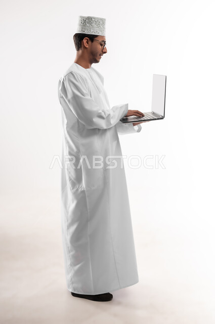 The concept of technological development and progress, the use of modern technical devices, a portrait of a young Gulf Arab Omani man wearing a dishdasha, a cloak and glasses using a laptop, completing work tasks remotely via a laptop, a full-length body image, a white background