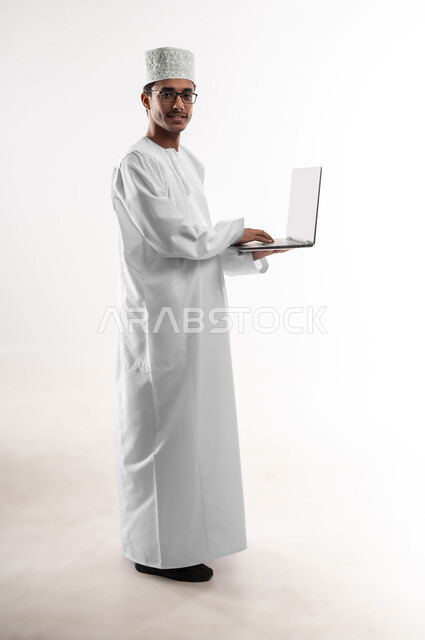 The concept of technological development and progress, the use of modern technical devices, a portrait of a young Arab Gulf Omani man wearing a dishdasha, a sleeve and medical glasses, looking at the camera and using a laptop, completing work tasks remotely via a laptop, a full-length body image, a white background