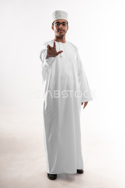 Teaching sign languages ​​to the deaf and dumb, interest in elegance and appearance, portrait of a young Gulf Arab Omani man wearing a dishdasha, a turban and glasses, looking at the camera, raising two fingers as the number two, communicating with people with disabilities audibly or vocally, full-length body image, white background