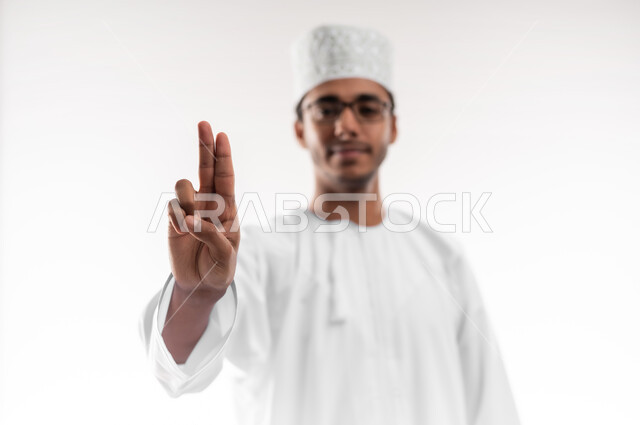The concept of request and permission, interest in elegance and appearance, portrait of a young Gulf Arab Omani man wearing a dishdasha, a sleeve and eyeglasses, raising two fingers up to indicate the number two, specifying a specific location, white background