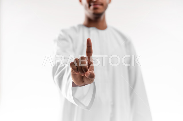 The concept of request and permission, interest in elegance and appearance, portrait of a young Arab Gulf Omani man wearing a dishdasha, raising his index finger up to indicate the number one, specifying a specific location, white background